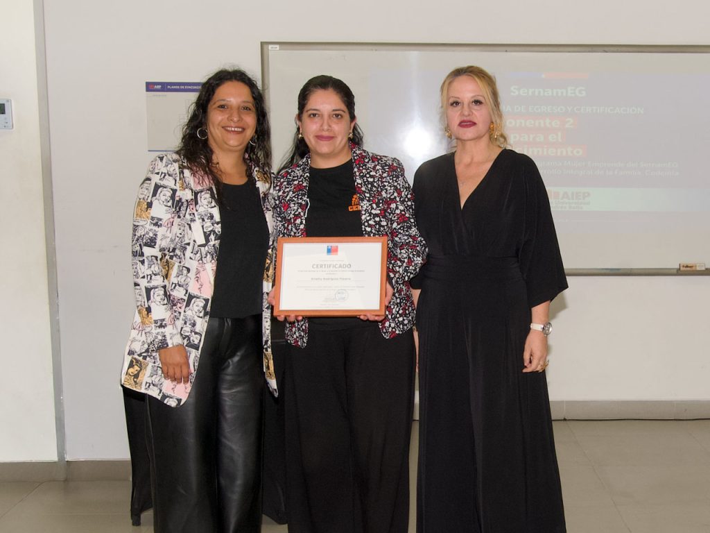 Entrega de diplomas en un aula de AIEP. Tres mujeres posan de pie; la del centro sostiene un certificado enmarcado de SernamEG, flanqueada por dos profesionales. Detrás se observa una pizarra con una proyección del evento.
