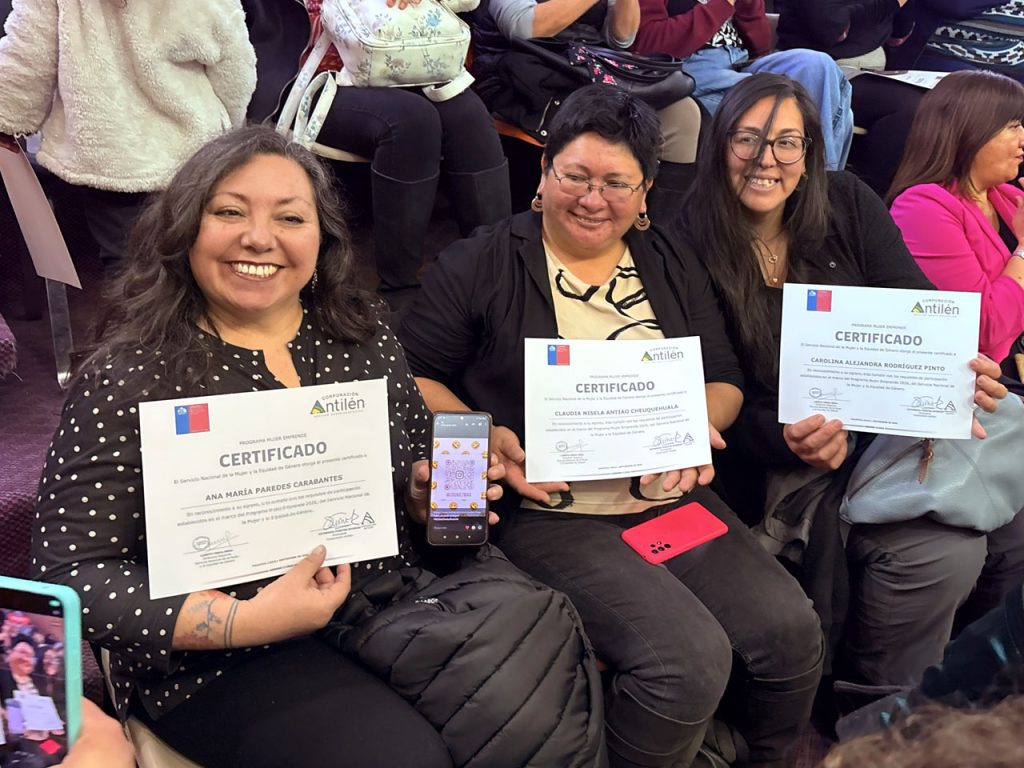 Tres mujeres sonrientes sentadas juntas muestran con orgullo sus certificados de la Corporación Antilén, emitidos en colaboración con el Servicio Nacional de la Mujer y la Equidad de Género de Chile.