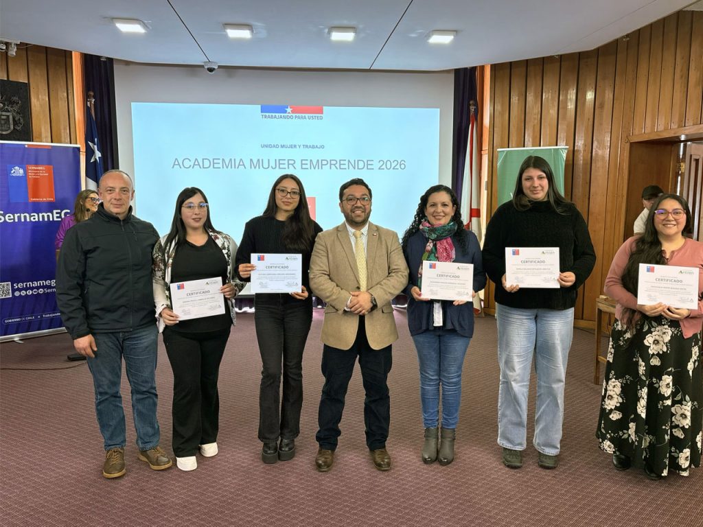Fotografía grupal sobre un escenario. Siete personas están alineadas de pie; cinco de ellas muestran certificados impresos de la Corporación Antilén y SernamEG. De fondo se observa una gran pantalla que proyecta el texto "Academia Mujer Emprende 2026 - Unidad Mujer y Trabajo". A la izquierda destaca un pendón corporativo de SernamEG del Gobierno de Chile.