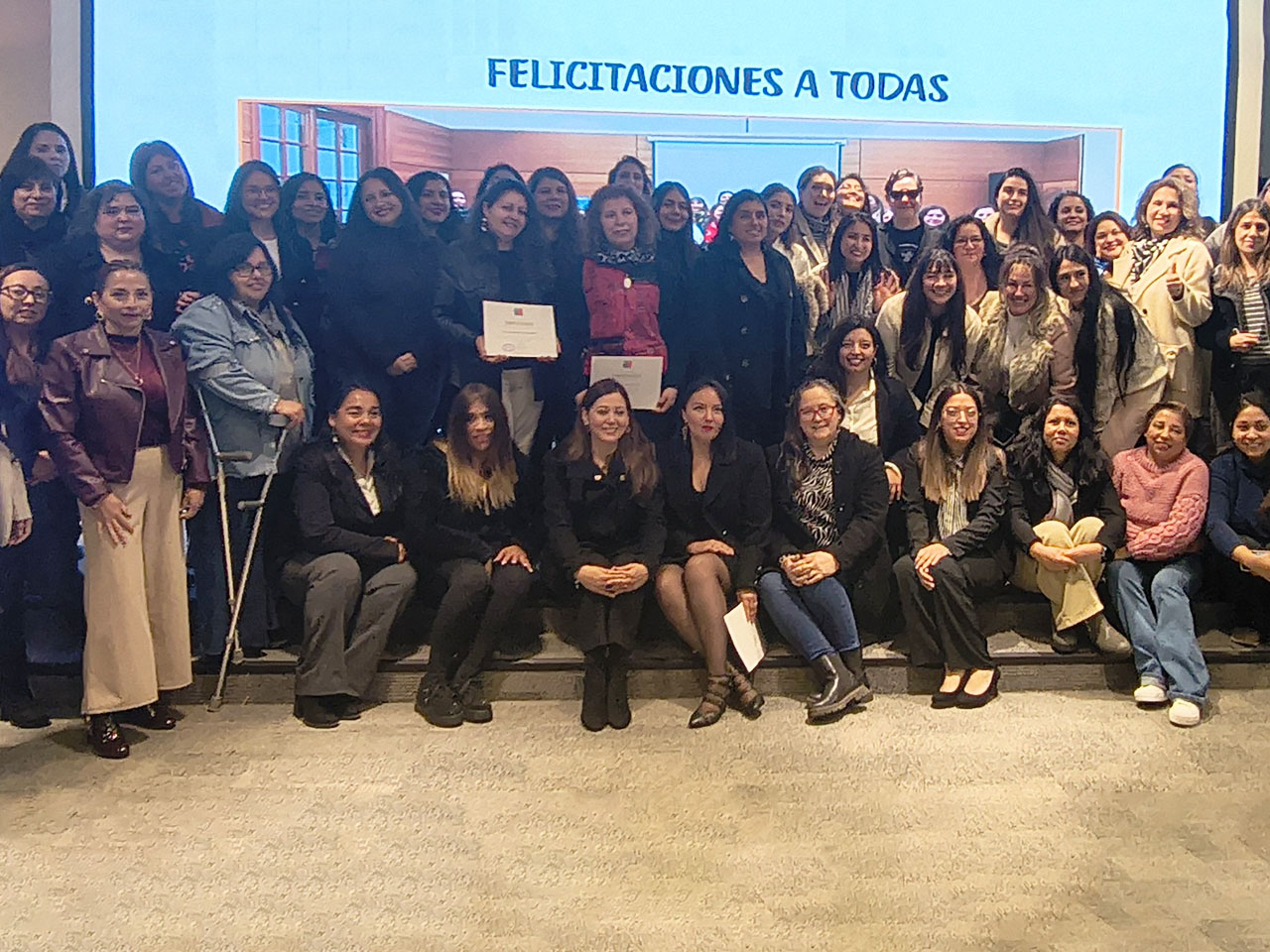 Un grupo numeroso de mujeres posa sonriente para una fotografía grupal en un auditorio o escenario. Dos de ellas sostienen diplomas de certificación en el centro. Al fondo, una pantalla digital proyecta una presentación con el título "FELICITACIONES A TODAS" en letras azules.