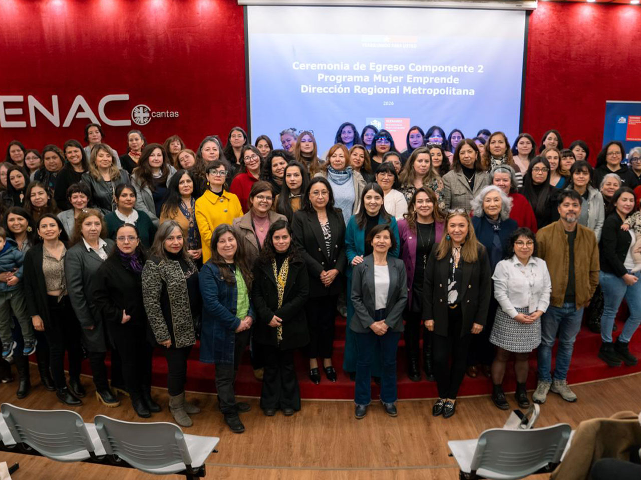 Un gran grupo de mujeres posa sonriente en un auditorio durante la Ceremonia de Egreso Componente 2 del Programa Mujer Emprende de la Dirección Regional Metropolitana del SernamEG, con el logo de ENAC Caritas visible al fondo