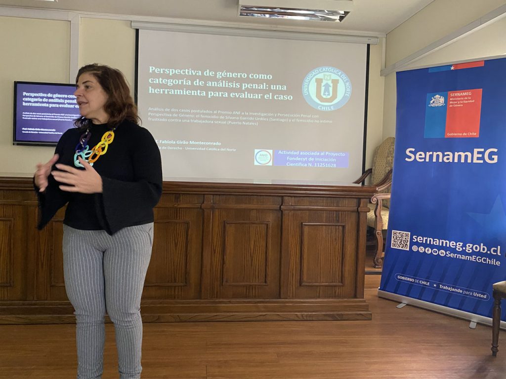 Una expositora realiza una presentación gesticulando con las manos en un aula, con una pantalla de fondo que detalla el proyecto Fondecyt sobre perspectiva de género en el análisis penal y un lienzo institucional de SernamEG a la derecha.