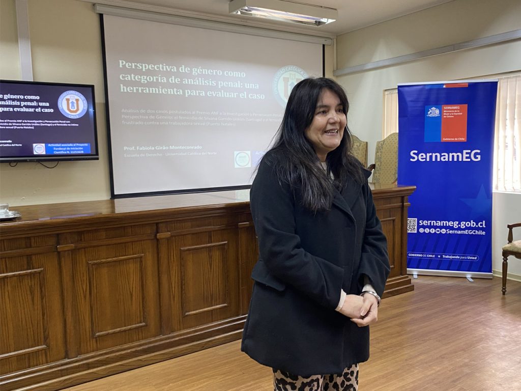 La profesora Fabiola Girão Monteconrado expone en un aula frente a una pantalla proyectada con el título "Perspectiva de género como categoría de análisis penal", junto a un lienzo institucional de SernamEG.