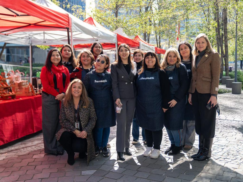 Fotografía grupal al aire libre en una feria institucional. Un grupo de mujeres, algunas con delantales oscuros con el logotipo de SernamEG, posan sonrientes junto a autoridades en un espacio público con carpas rojas y stands de fondo.